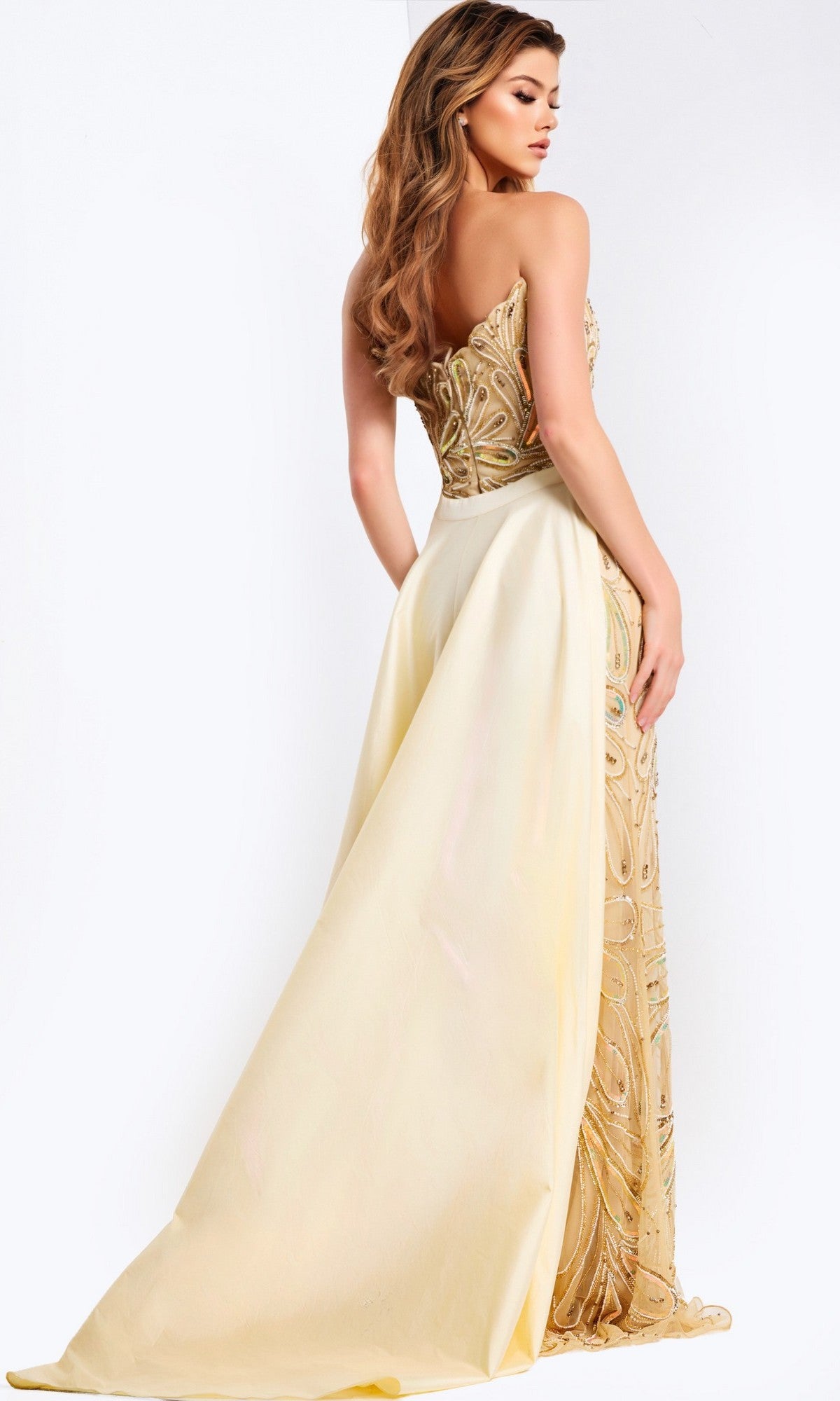A woman in the Jovani 49255 long prom dress, featuring a strapless gold-embellished beaded bodice and an attached cream train, stands sideways and glances over her shoulder.